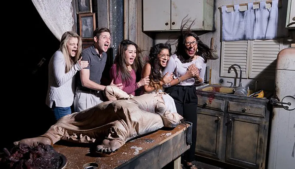 A group of people appear terrified and scream while gathered around a table with a fake gory figure as part of what looks like a haunted house or horror-themed attraction