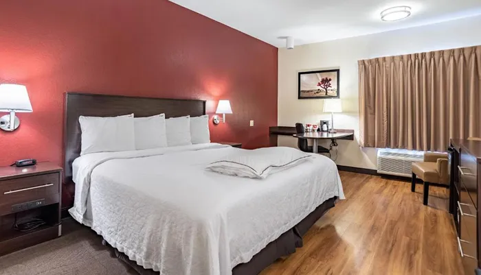 A neatly arranged hotel room with a queen-sized bed a red accent wall and a small sitting area next to the window