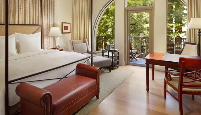 The image shows a well-appointed hotel room with a large bed elegant furniture and French doors leading to a balcony with a view of trees