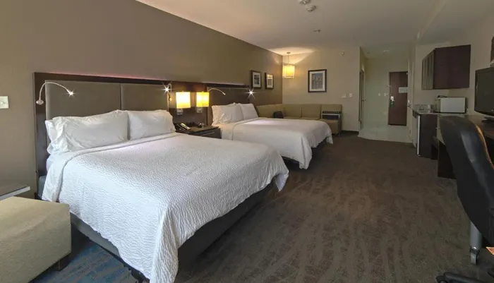 This is an image of a tidy furnished hotel room featuring two beds with white linens a seating area and an office desk