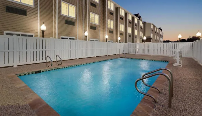 This is an image of an outdoor swimming pool enclosed by a white fence with apartment buildings in the background taken in the evening as the lights are on