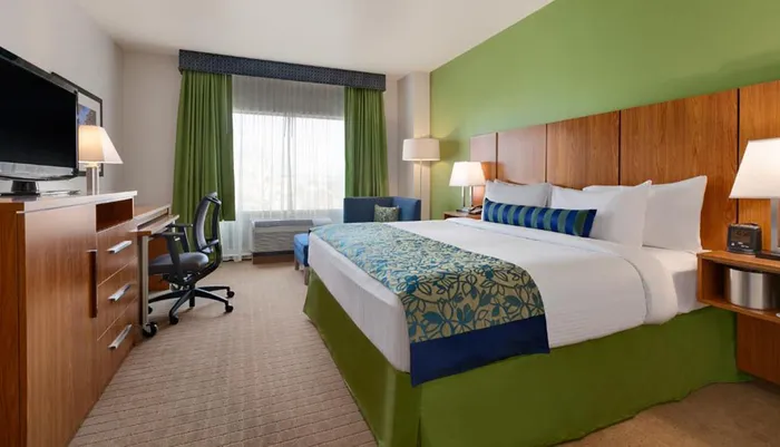 The image shows a neatly arranged hotel room with a king-size bed a desk with a chair and a flat-screen TV