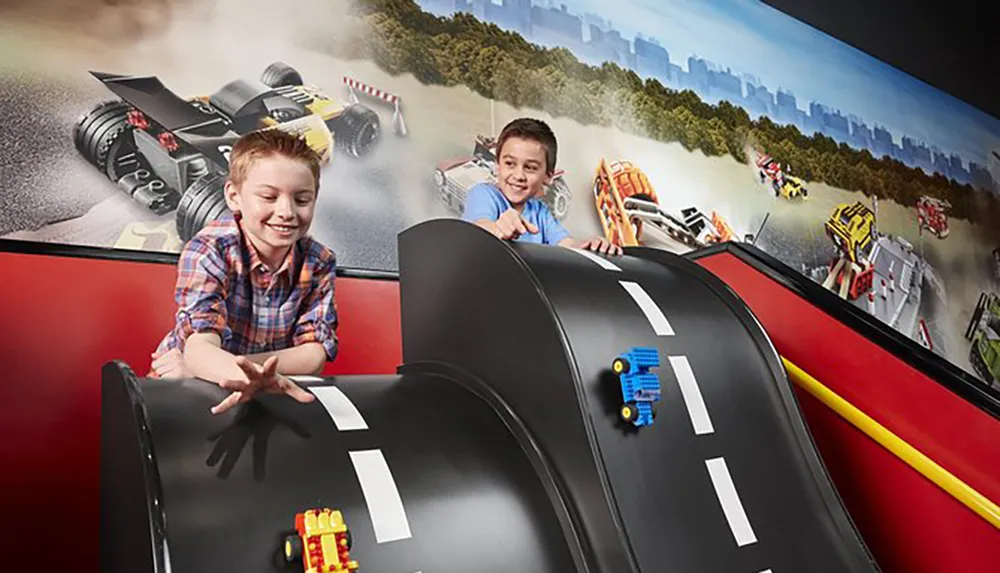 Two children are joyfully playing with toy cars on a specially designed race track that emulates a real racing scenario with a dynamic racing-themed mural in the background