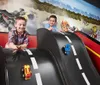 Two children are smiling and playing with toy cars on a curved oversized race track model with a dynamic scene of racing cars in the background