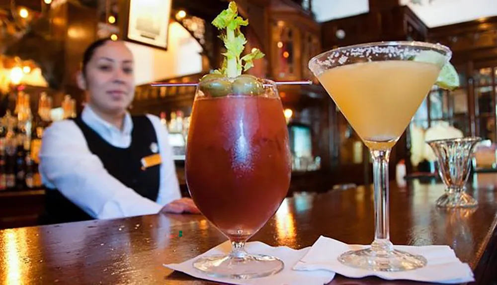 A bartender stands behind a bar counter where two beverages a Bloody Mary and a martini are prominently displayed in the foreground