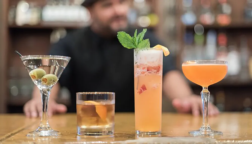 The image shows a selection of four different cocktails on a bar surface with a blurry bartender in the background