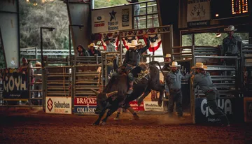 A rodeo rider is mounted on a bucking bull inside an arena, surrounded by spectators and rodeo personnel.