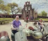 Two people are happily posing on Vespa scooters with sidecars in front of a historic stone mission building under a bright blue sky