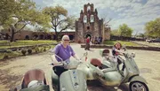 Two people are happily posing on Vespa scooters with sidecars in front of a historic stone mission building under a bright blue sky.