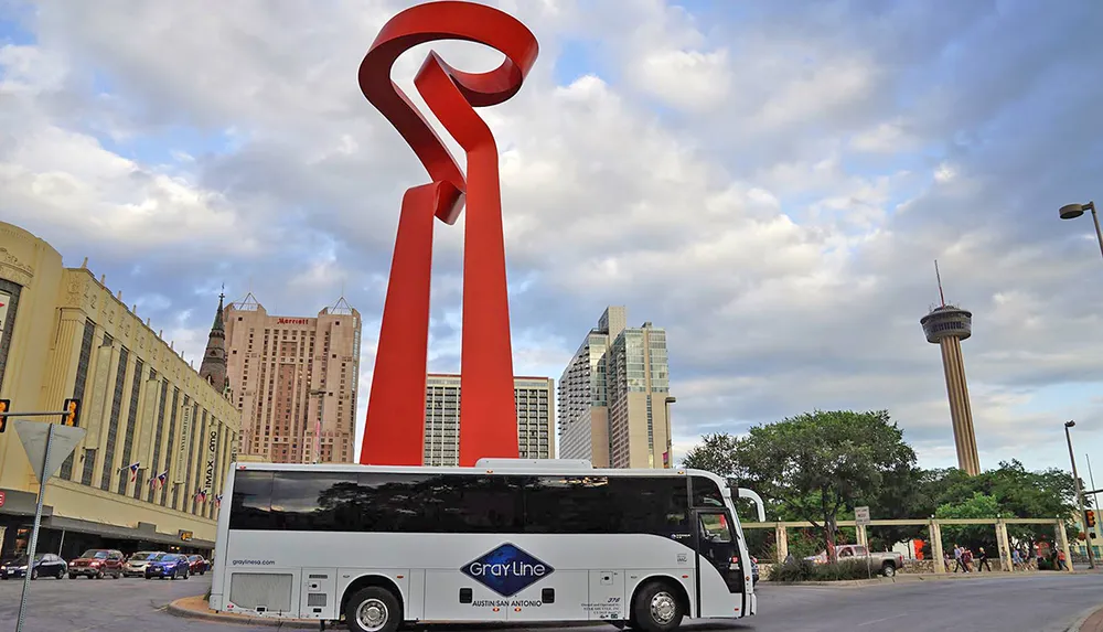 A tour bus drives through an urban environment featuring a distinctive red sculpture and a tower in the background under a partly cloudy sky