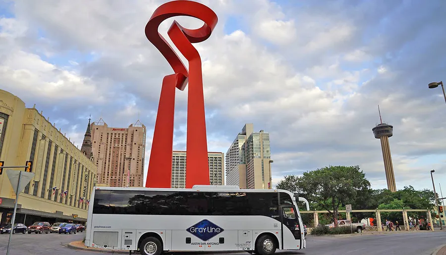A tour bus drives through an urban environment featuring a distinctive red sculpture and a tower in the background under a partly cloudy sky.