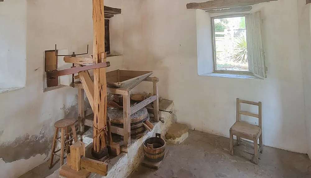 The image shows a rustic old-fashioned room featuring a traditional grain mill with wooden gears and a grinding stone a simple wooden chair and a bucket illuminated by natural light from a window with a view of outdoor greenery