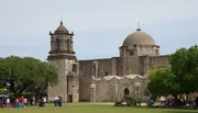 The image shows visitors around an old stone mission with a distinctive dome and bell tower, surrounded by trees under a clear sky.