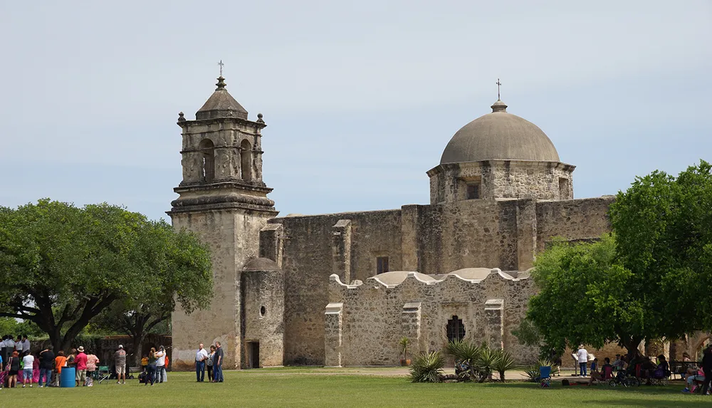 The image shows visitors around an old stone mission with a distinctive dome and bell tower surrounded by trees under a clear sky