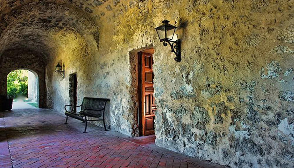 The image shows a serene arched passageway with textured walls a wooden door a lantern and a bench evoking a sense of historical charm