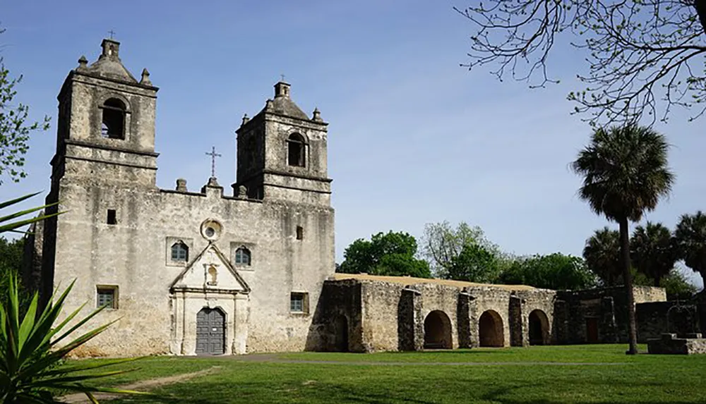 The image shows an old weathered mission with dual bell towers and a surrounding wall against a backdrop of trees under a clear blue sky