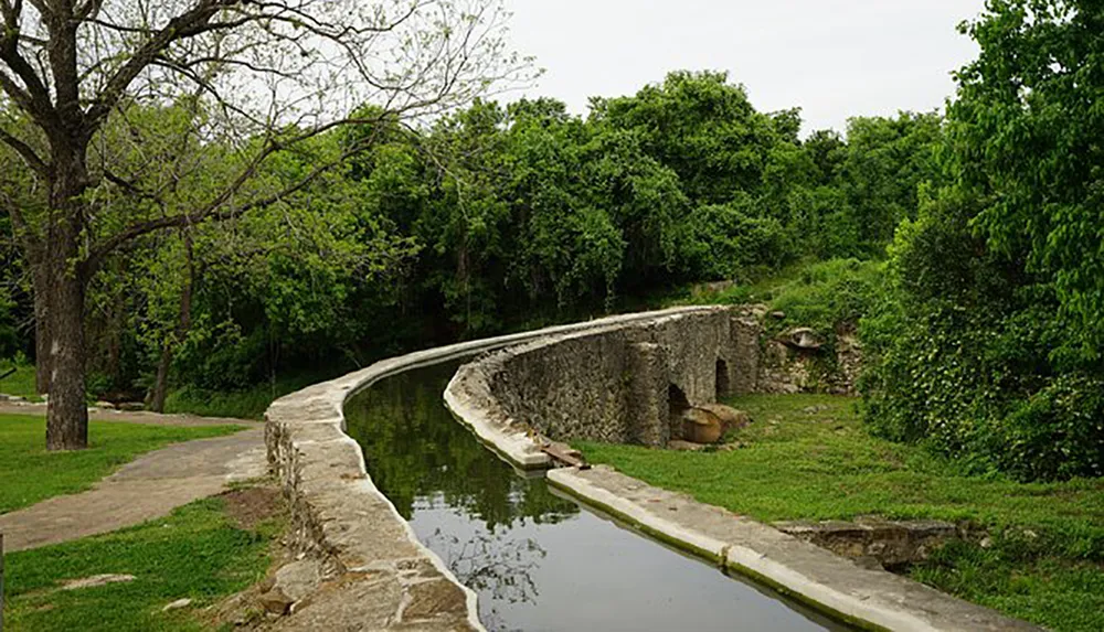 The image shows a tranquil park scene with a curving water canal bordered by a stone wall and a pathway surrounded by abundant greenery and trees