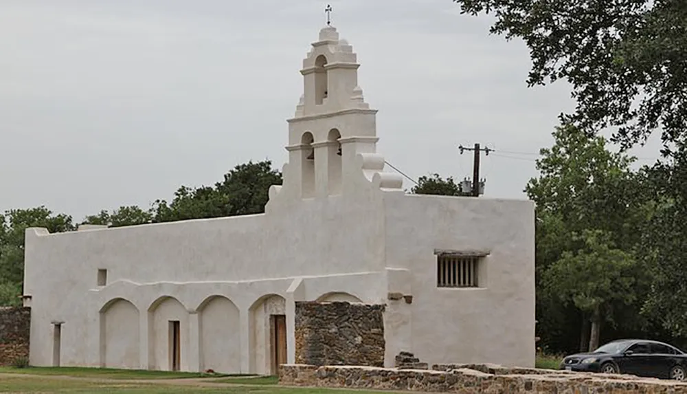 The image shows a white historic-looking mission-style church with a bell tower arched doorways and a stone foundation set against a backdrop of green trees and an overcast sky