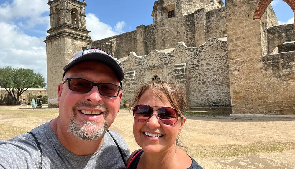 Two smiling individuals are taking a selfie in front of an ancient stone building with a large bell tower under a clear sky