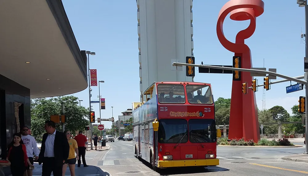 A red double-decker sightseeing bus is passing through an intersection with a large red sculpture in the background