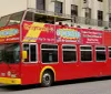 A red double-decker sightseeing tour bus is parked on the street advertising hop-on hop-off services and air conditioning with people seated on the upper open-air level