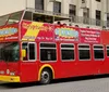 A red double-decker sightseeing tour bus is parked on the street advertising hop-on hop-off services and air conditioning with people seated on the upper open-air level