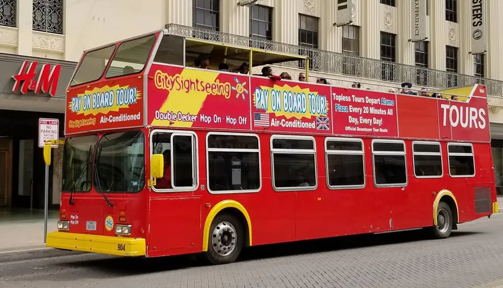 A red double-decker sightseeing tour bus is parked on the street advertising hop-on hop-off services and air conditioning with people seated on the upper open-air level