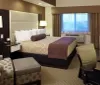 The image shows a neatly arranged hotel room with a king-size bed patterned chairs a work desk and a flat-screen TV