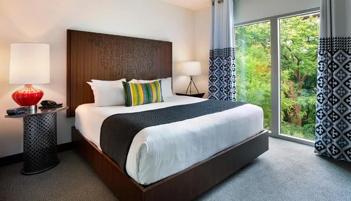 The image shows a neatly arranged modern hotel room with a large bed stylish curtains a red lamp and a view of greenery outside the window
