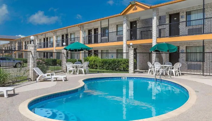 This image shows a sunny outdoor scene with a clear blue swimming pool in the foreground and a two-story motel with balcony access rooms in the background complete with umbrellas and seating arrangements around the pool area