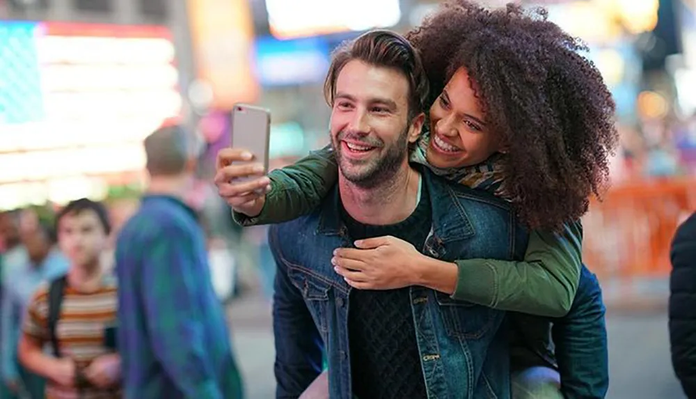 A joyful couple is taking a selfie together amidst a bustling city scene with colorful lights in the background