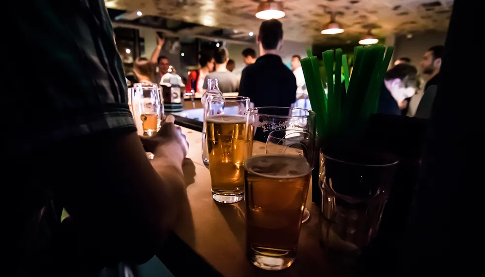 The image captures a dimly lit social scene with a focus on several glasses of beer on a counter in the foreground of a bustling bar atmosphere