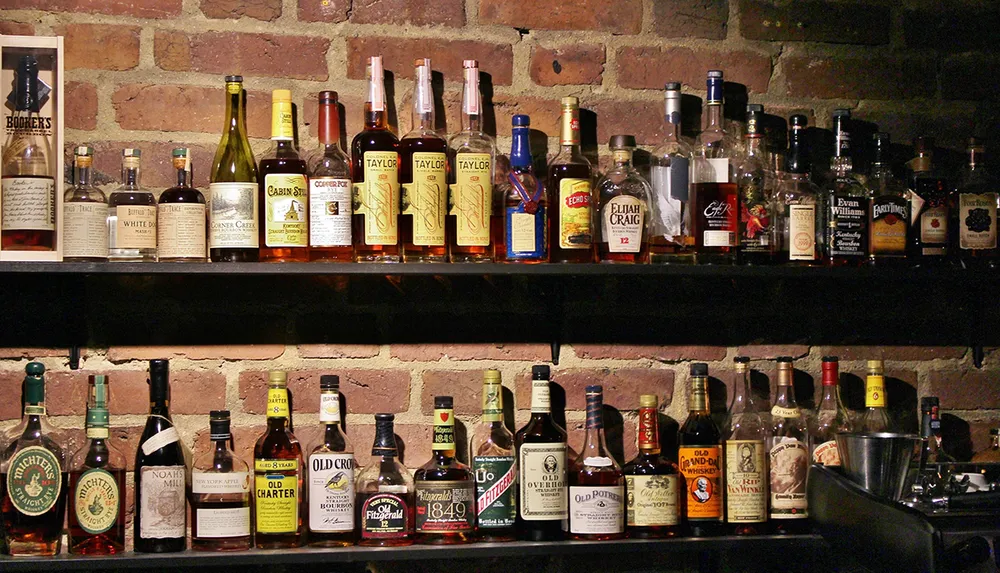 A diverse collection of liquor bottles is displayed on shelves against a brick wall likely in a bar or restaurant setting