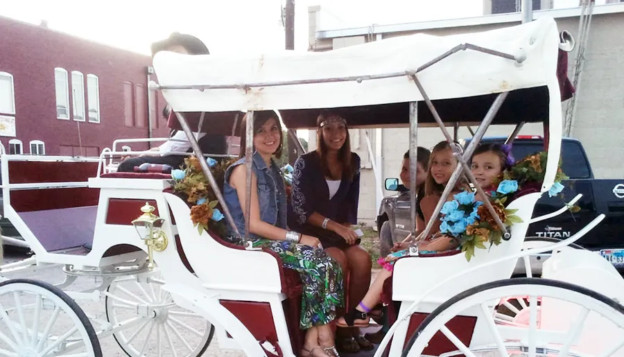 Four passengers are smiling while seated in a decorated horse-drawn carriage on an urban street.