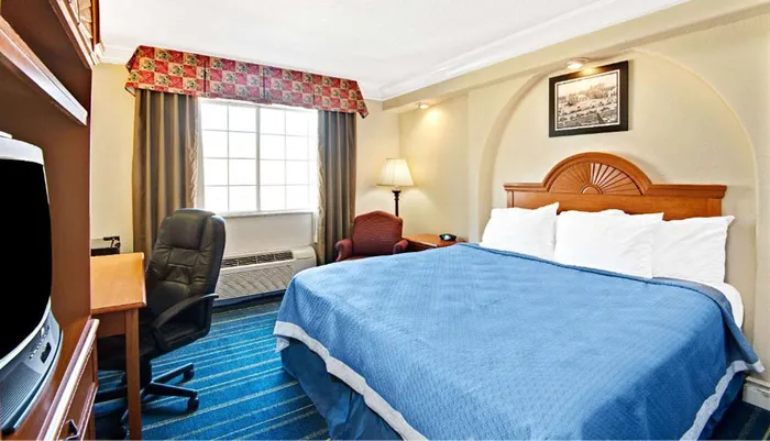 The image shows a neatly arranged hotel room with a large bed an office chair and desk a window with curtains and a wall-mounted picture