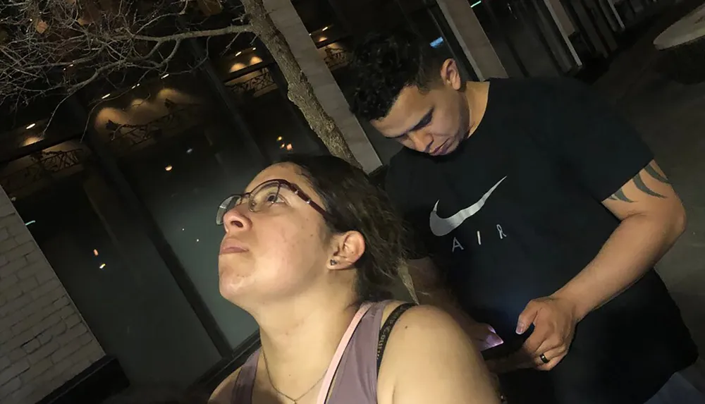 A woman and a man appear to be waiting or observing something standing outdoors at night with a tree and building in the background