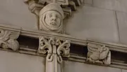 The image shows an architectural detail featuring a carved stone face framed by decorative elements, including winged figures, on a building's façade.