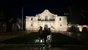Three people are posing for a photo at night in front of the historic Alamo mission in San Antonio, Texas, illuminated by spotlights.