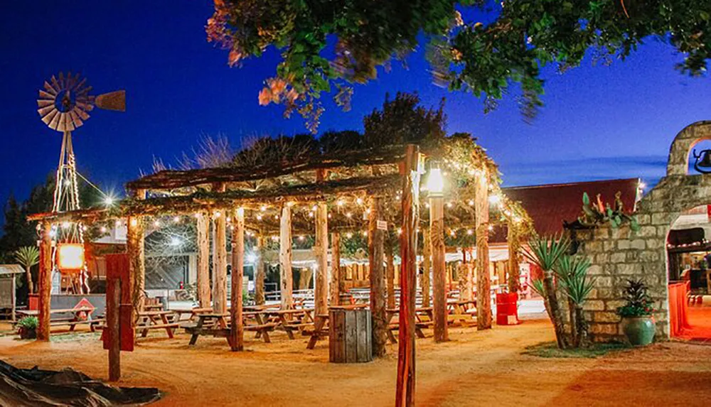 The image shows a rustic outdoor dining area adorned with twinkling string lights and a windmill in the background evoking a cozy country atmosphere