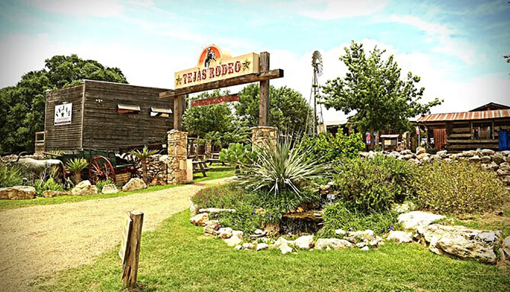 The image shows a rustic entrance to a Texas rodeo with a wooden structure a sign featuring a longhorn skull and a garden with native plants and a wagon wheel