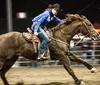 A focused rider in a blue shirt and cowboy hat is galloping on a brown horse in an equestrian arena