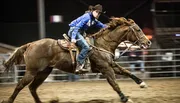 A focused rider in a blue shirt and cowboy hat is galloping on a brown horse in an equestrian arena.