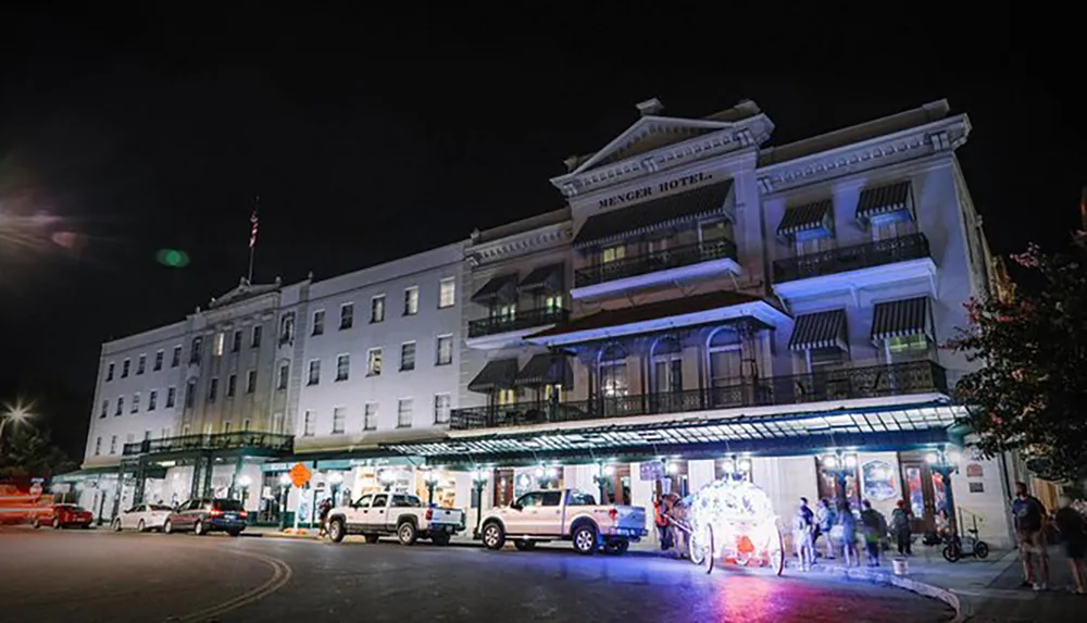 The image shows the Menger Hotel at night illuminated by street lights with a horse-drawn carriage in front adding to the historical ambiance of the scene