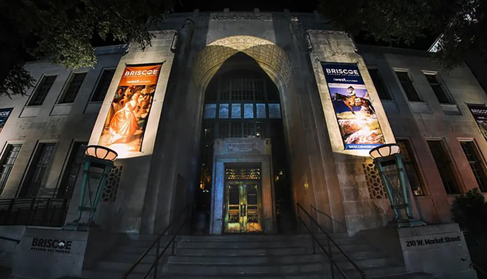 The image shows the illuminated facade of the Briscoe Western Art Museum at night featuring grand arches and large banners displaying artwork
