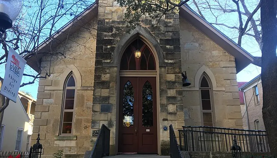 The image shows the front facade of a quaint stone church with a Gothic arched doorway, framed by trees under a clear sky.