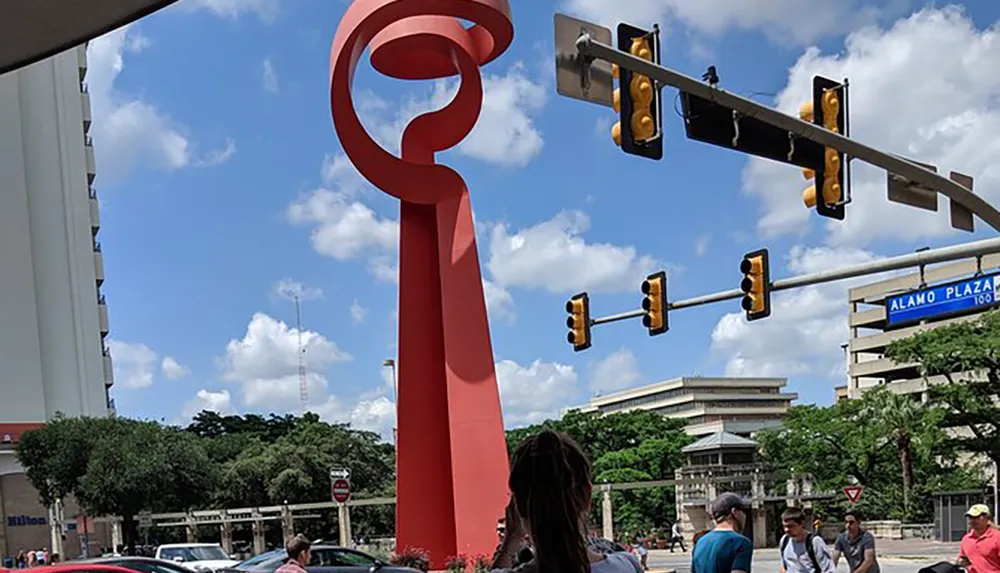 The image captures an urban scene featuring pedestrians traffic lights and a large red abstract sculpture under a blue sky with fluffy clouds