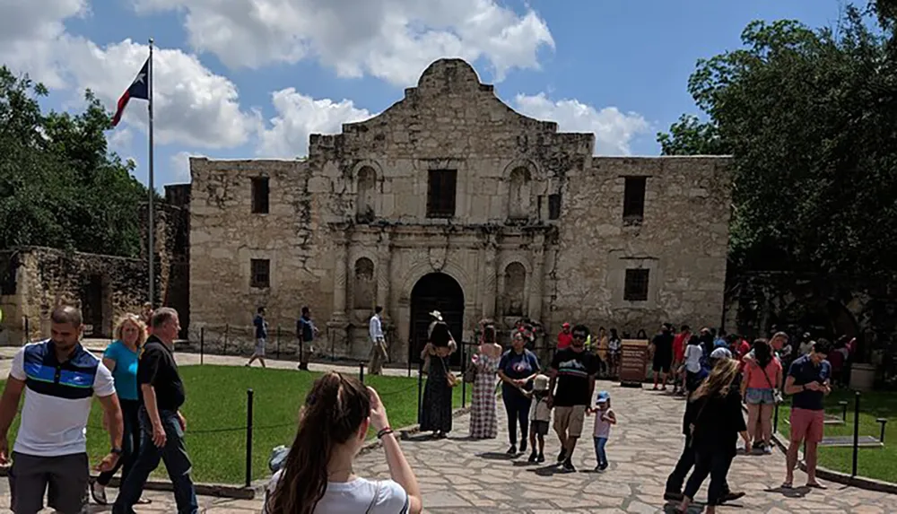 Visitors are gathered around the historic Alamo mission on a sunny day with several people taking photos and a Texas flag flying in the background