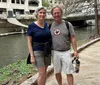 A smiling couple is posing for a photo while standing by an urban riverwalk with a bridge and some ducks in the background