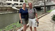A smiling couple is posing for a photo while standing by an urban riverwalk with a bridge and some ducks in the background.