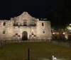Four individuals are posing for a photo in the evening in front of the historic Alamo mission building